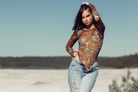 Slim young woman in ornamental bodysuit touching hair while resting on sandy beach on sunny day