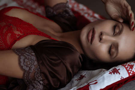 Side view of sexual young female with long dark hair in elegant red bra lying on comfortable bed with closed eyes near decorated Christmas treeの写真素材