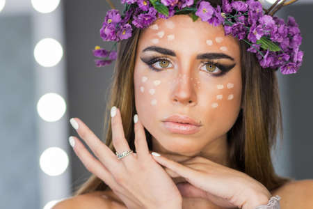 Young woman with violet flower wreath branch horns and deer spots on face standing looking at camera...