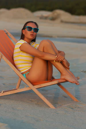 Full body alluring woman in sexy shirt and hat sitting sensually on sandy beach in sunset light