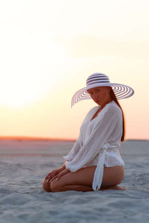 Full body alluring woman in sexy shirt and hat sitting sensually on sandy beach in sunset lightの写真素材
