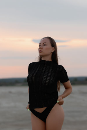 Alluring young brunette wearing black transparent t shirt with sexy panties standing on sandy shore in twilightの写真素材