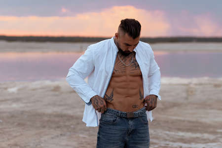 Muscular athletic sexy man in white shirt with bare-chested rests on beach. Portrait of tanned fit male model on vacation.の写真素材