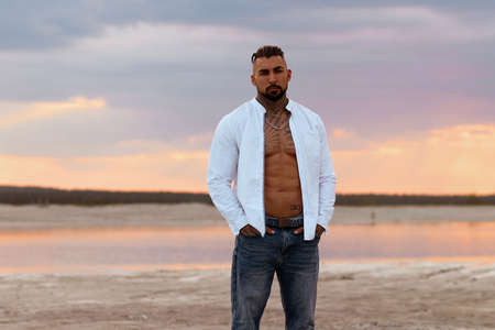 Muscular athletic sexy man in white shirt with bare-chested rests on beach. Portrait of tanned fit male model on vacation.の写真素材