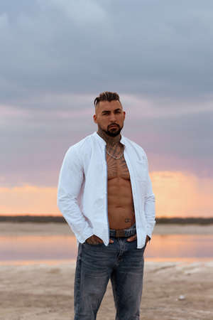 Muscular athletic sexy man in white shirt with bare-chested rests on beach. Portrait of tanned fit male model on vacation.の写真素材