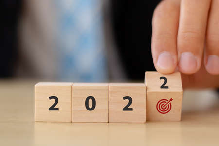 Business goals in 2022 concept. Businessman hand flips wooden cubes "2022" and "dart board has dart arrow hitting" icon on beautiful  background and copy space. For starting business annual plan.の写真素材
