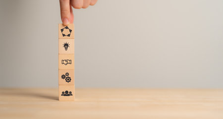 Business Synergy Concept. Building business success.A hand balancing wooden blocks with icons representing teamwork, innovation, collaboration, and operations, symbolizing business synergy.の写真素材