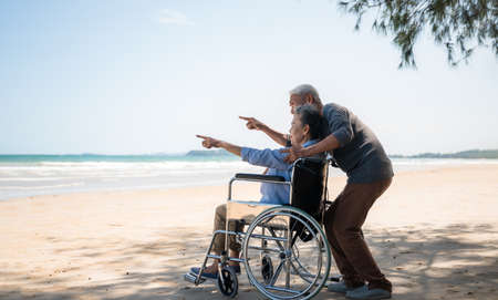 Side view of asian senior couple relaxing smile and enjoying with pointing and look to sea on beach under tree. Happy couple elderly retire resting and relax wife is disabled and sitting on wheelchairの写真素材