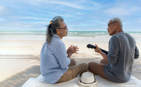 Asian senior couple tourist wear sunglasses is clap fun and sitting singing with playing ukulele on beach in summer vacation. Happy family elderly older retire relax lifestyle together on seaside.の写真素材