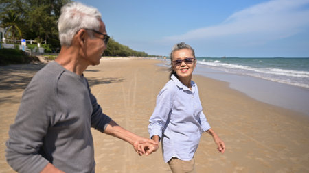 Asian senior woman wear sunglasses smile and holding hand husband walking running on beach in summer vacation. Happy family couple hair gray elderly older retirement relax together on seaside outside.の写真素材