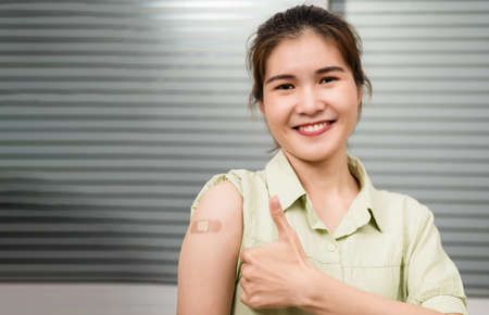 Happy Asian woman sitting on couch smile and giving thumb up after getting vaccination immunity for protectionの写真素材