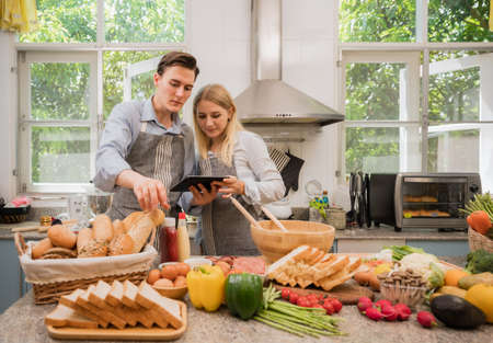 Young couple in apron happy and fun cooking salad together with digital tablet at kitchen in home....