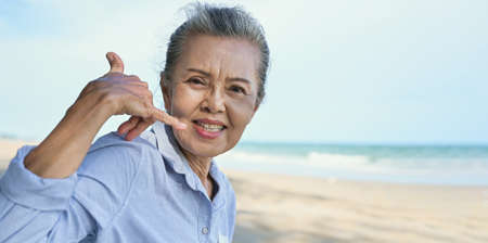 Asian senior woman sitting smiling and using hand sign is phone with talking on sea beach in summer...