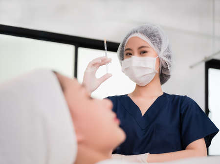 Cosmetologist wear face mask and gloves with holding injection syringe for preparing to face augmentation for anti wrinkle in beauty clinic. Asian beautician doing anti aging procedure in beauty room.の写真素材