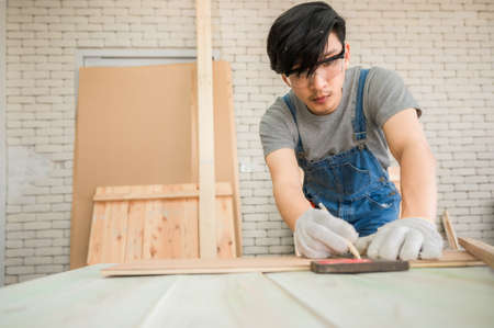 Young man carpenter wearing safety goggle using pencil and Iron ruler draw line on plank wood for furniture manufacturing in workplace. Concept of asian carpenter measuring and marking on wooden plankの写真素材