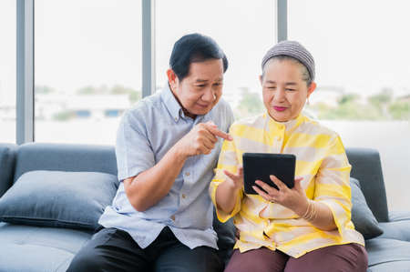 Happy elderly asian couple sitting on couch and using digital tablet computer for online shopping...
