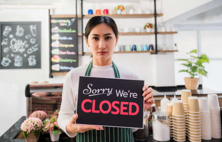Unhappy barista woman in apron and holding closed shop signboard in cafe. Portrait of sorry asian...