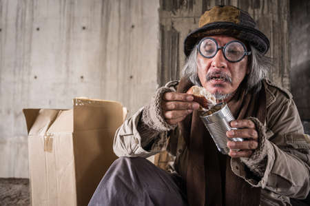 Happy homeless old man sitting eating bread and canned food from donation of kind people. Asian poor...