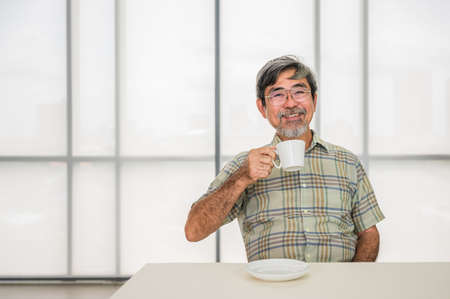 Happy senior asian man sitting on chair and holding coffee cup ready to drink in living room. Elderly man gray hair beard and smile with coffee at table in morning. Lifestyle after retirement concept.の写真素材