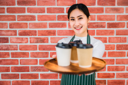 Beautiful asian young barista woman in apron holding tray coffee with serving to customer in cafe. Portrait of girl barista smiling and showing wooden tray with coffee glass on brick wall background.の写真素材