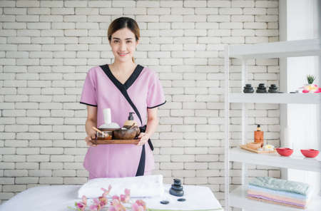 Happy young asian woman in uniform standing and holding tray of spa treatment accessories in spa massage room. Massage therapist female smile and welcome with service wellness for high quality.の写真素材