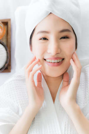 Beautiful asian young woman touching her face by hands after clean fresh skin on face. Happy lady in white dress with lying down and enjoy of skin care treatment in spa salon. Skincare and spa conceptの写真素材