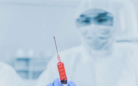 Male medical scientist researcher in protect suit and hygiene mask standing with holding syringe and blood plasma to vaccine research for protective infection coronavirus or covid-19 in laboratory.の写真素材