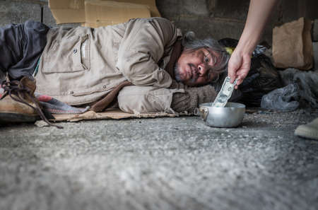Homeless senior man with gray beard and grey hair with wearing sweater and blanket sleeping the sidewalk and street walkway. Hand of kind woman giving banknote and donate money to poor man beggar.の写真素材