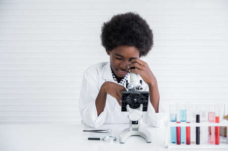 Happy african child boy student is learning and test science chemical with colorful liquid in tube by looking from microscope in laboratory room at to school. Education and science research concept.の写真素材