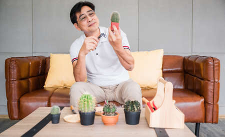 Asian elderly retired man sitting on couch is planting and decoration cactus in living room at to home. Lifestyle and hobby concept of retirement aged senior male.の写真素材