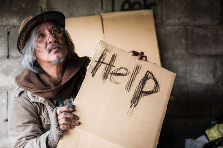 Old homeless man with gray beard sitting hungry and food beggar with holding cardboard sign seeking help from people walking on street. Closeup and focus to word Help on cardboard sign. Hope concept.の素材 [FY310174624642]