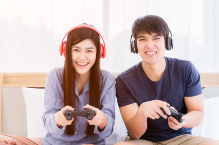 Happy young couple wear headphone and holding joystick playing video game on console to fun. Asian couple wearing earphone and sitting with relaxation by technology playstation on bed in bedroom.の写真素材