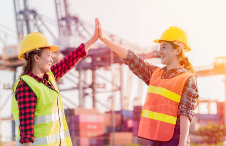 Two asian staff woman in yellow safety helmet with standing and putting hands up for job successful after from control ship loading cranes. Happy team putting hands up and show the unity.の写真素材