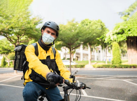 Portrait of Asian food delivery rider man in uniform wear medical face mask and helmet use bicycle...