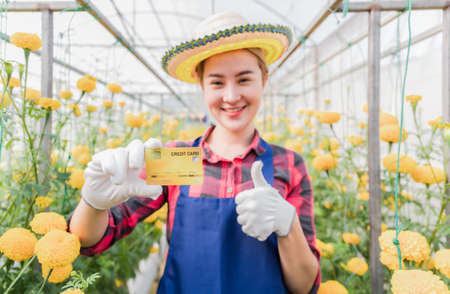 Happy asian woman standing and holding credit card in yellow marigold garden. Business owner and entrepreneur startup concept of female farmer in organic greenhouse.の写真素材