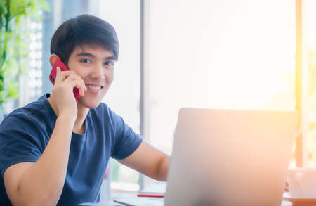 Happy asian man sitting talking by smartphone and working at home by laptop in home office. Student young handsome is calling to friend and learning online by notebook computer in living room.の写真素材