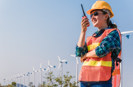 Woman engineer in uniform and yellow safety helmet using radio communication for staff contact to check wind turbine power in construction site renewable energy. Clean energy and environment concept.の写真素材