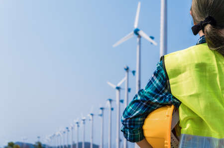 Woman engineer in uniform and holding yellow safety helmet with standing and checking wind turbine power in construction site renewable energy. Clean energy and environment concept.の写真素材