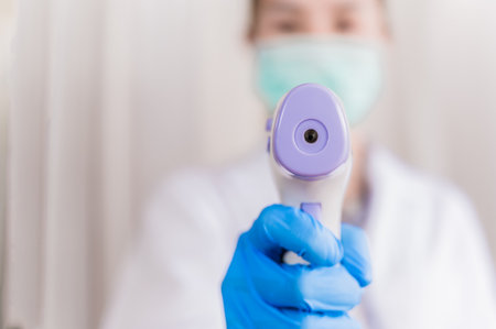 Closeup of infrared forehead thermometer gun. Woman doctor wear uniform and mask holding infrared...