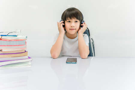 Child boy wear headphone and enjoy listening music by mobile phone with sitting on chair in classroom and book placed on white table for relax while break time. Back to school for concept.の写真素材