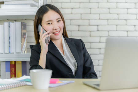 Portrait of asian business woman in suit smiling sitting on chair and talking by smartphone with...