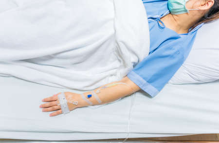 Young patient asian woman wear mask and lying on bed in hospital with IV saline drip to back of the hand. Virus or  concept.の写真素材