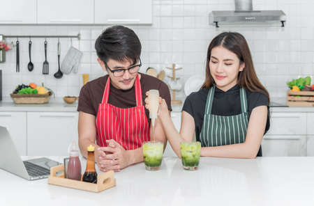 Asian couples opening laptop are preparing healthy breakfast by young female pouring milk into the tea glass and young man looking with happiness in kitchen room at to home. Food and drink healthy concept.の写真素材