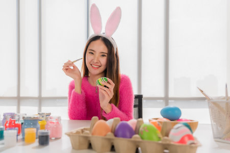Attractive asian young woman wearing bunny ears sitting smiling and painted easter egg with happy...
