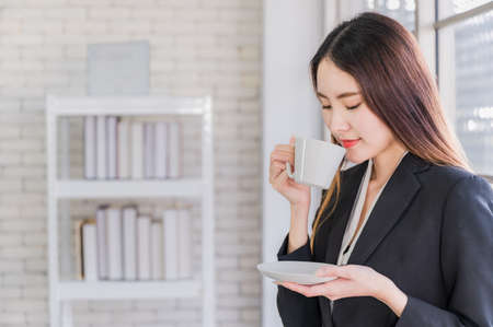 Beautiful young asian businesswoman in suit standing holding white coffee cup in office while having a coffee break.の写真素材