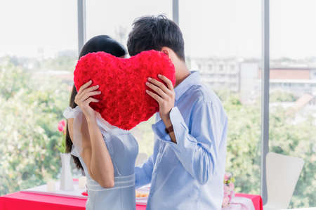 Side view of couple hiding behind red heart shape while standing in restaurant room on building. Valentines day and lover people concept.の写真素材