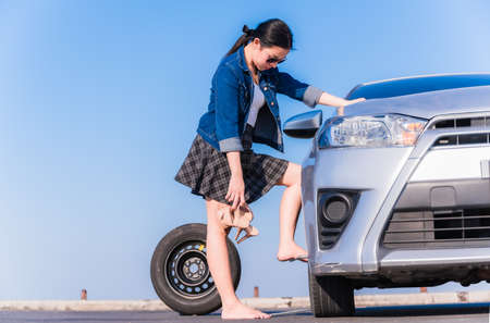 Beautiful asian woman treading on wrench while changing flat tire wheel on the side road and hand holding high shoes.の写真素材