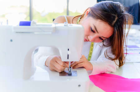 Beautiful asian woman tailor and seamstress sews input thread by hand into sewing machine. Designer...