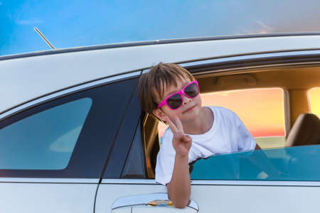 Happy boy wear sunglasses sitting in white car and show hand from making sign of victory by fingers on out of the car window while trip travel in holiday.の写真素材