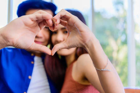 Closeup of asian young couple sitting on couch and looking through a heart shape made with their fingers to happily in living room at home.の写真素材